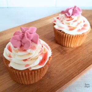 Two vegan vanilla cupcakes topped with a buttercream swirl and a pink royal icing flower.