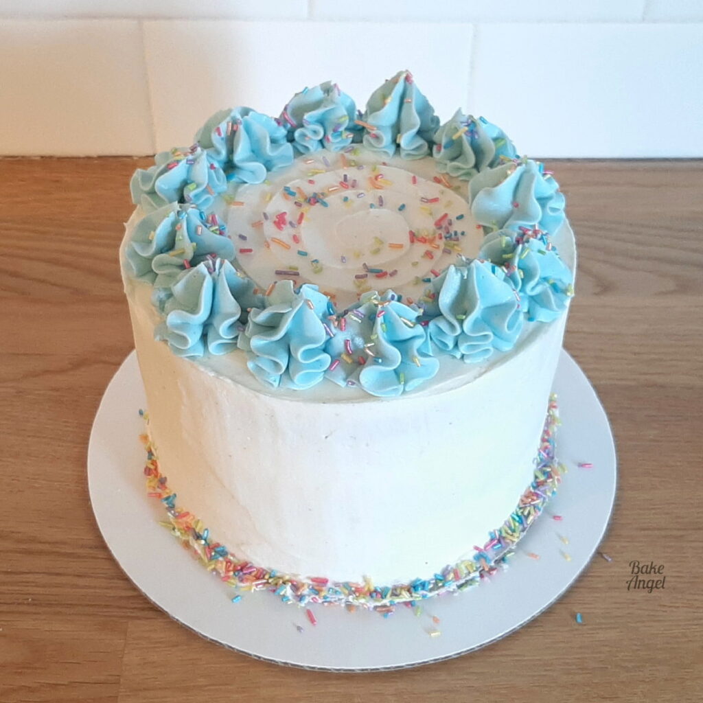 Vegan Vanilla Layer Cake coated in white buttercream with blue swirls on top and rainbow sprinkles. 