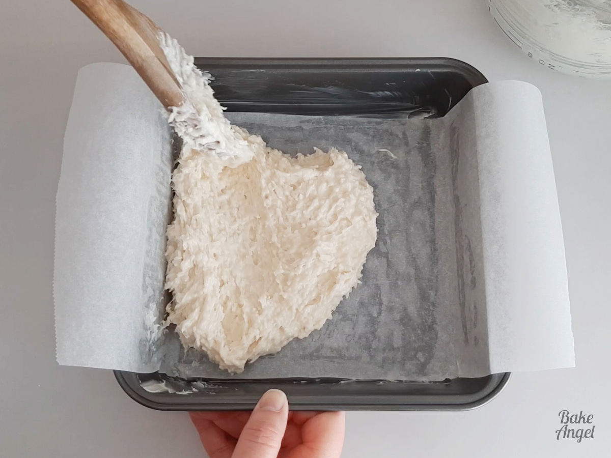 Using a wooden spoon to add white coconut ice mixture to a lined baking pan. 
