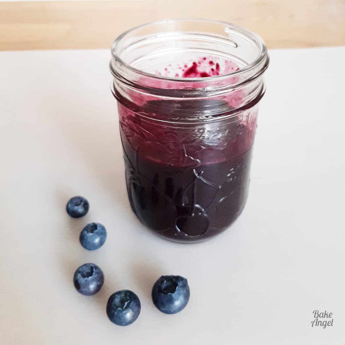 A glass mason jar filled with blueberry compote with 5 blueberries next to it on a white counter.