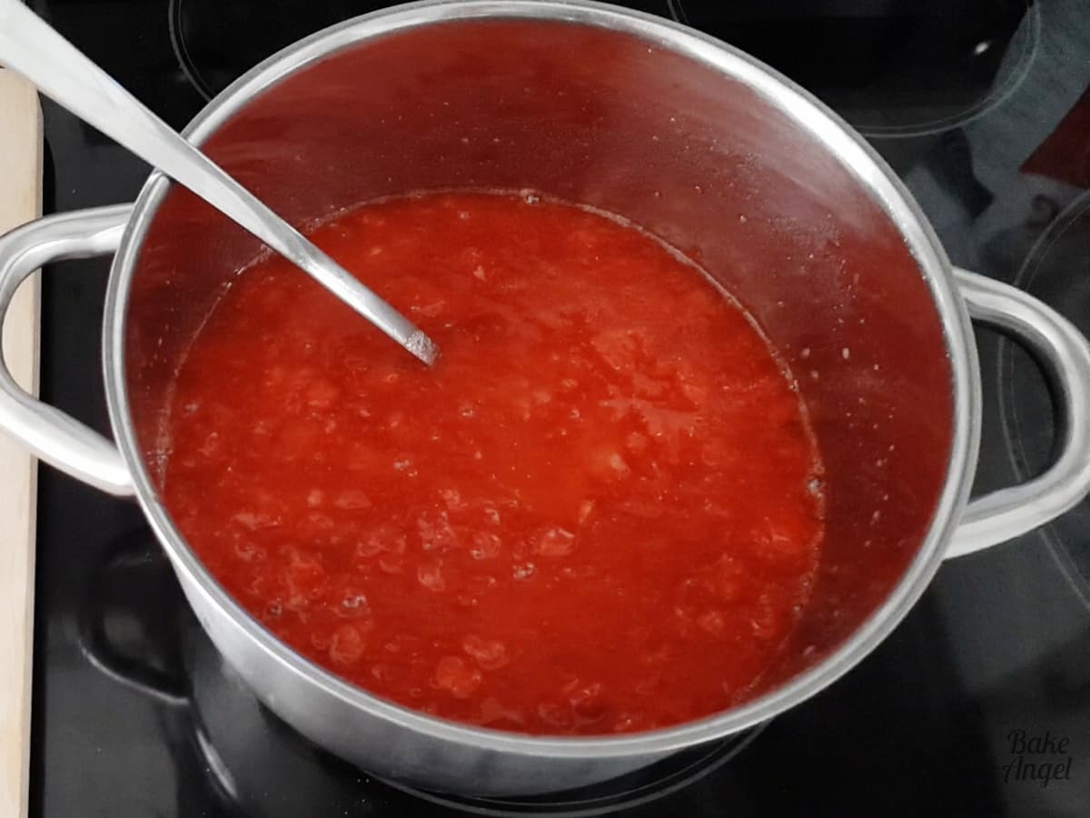 A large saucepan of strawberry jam ixture on a black stove with a silver ladle in the pan. 