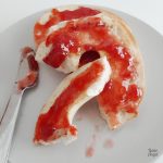 Close up of strawberry jam on a bagel on a plate with a knife on the left side.
