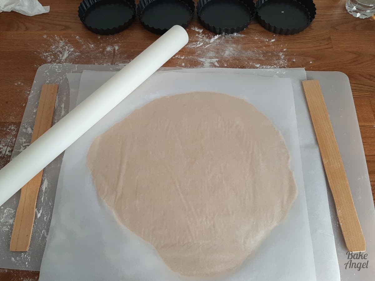 Rolling out pastry dough between parchment paper. A white rolling pin, 4 mini tart cases and two paint stirrer sticks are shown near the parchment paper. 