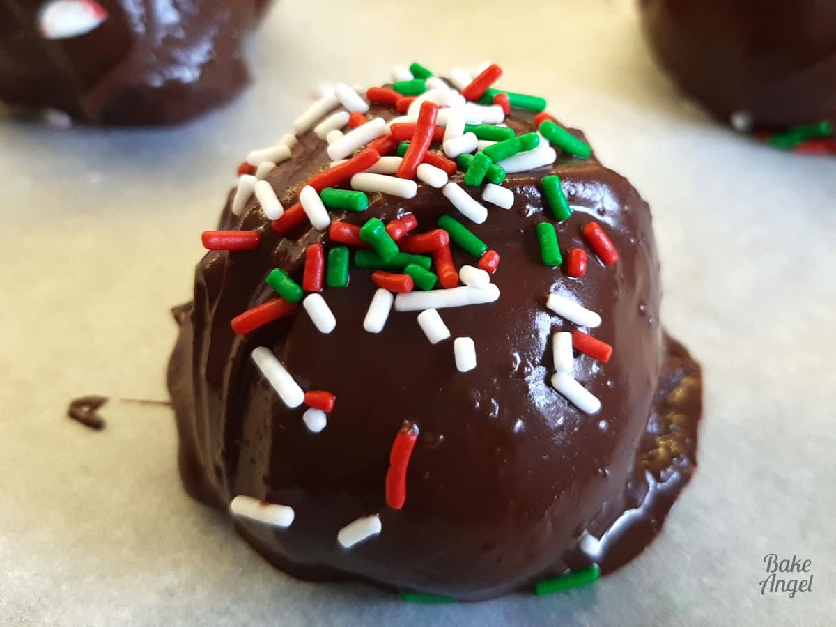Close up of a festive cake ball coated in chocolate with white, green and red sprinkles on top. 