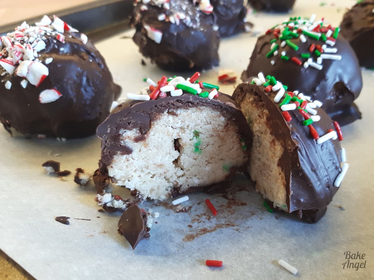 Close up of a festive cake ball cut in half showing the sprinkles inside and coated in chocolate with white, green and red sprinkles on top. 