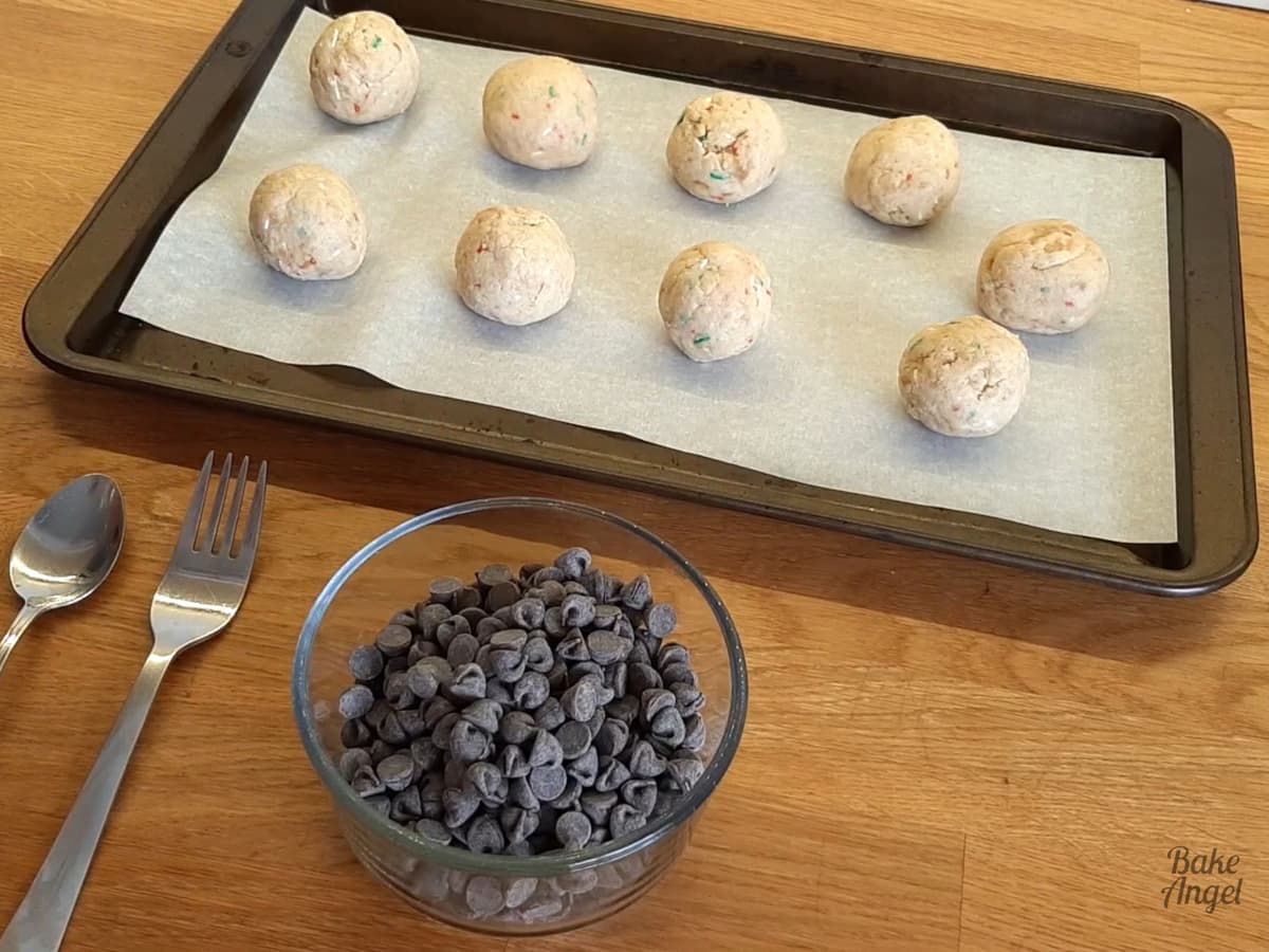 9 festive cake balls on a lined baking tray with a bowl of chocolate chips in front. 