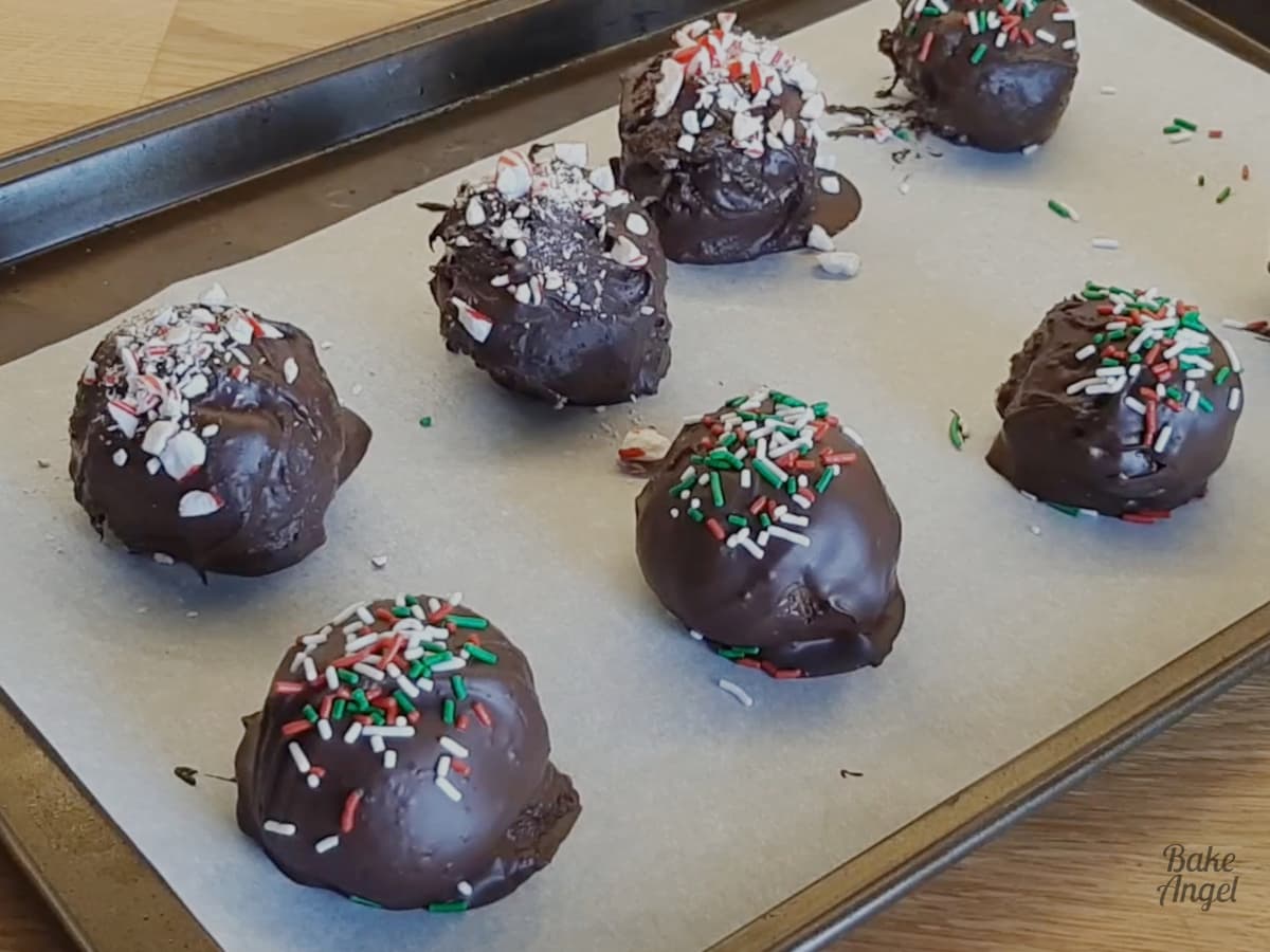 Decorated cake balls on a baking tray. 3 have red, white and green sprinkles on top and 3 have crushed candy canes on top. 