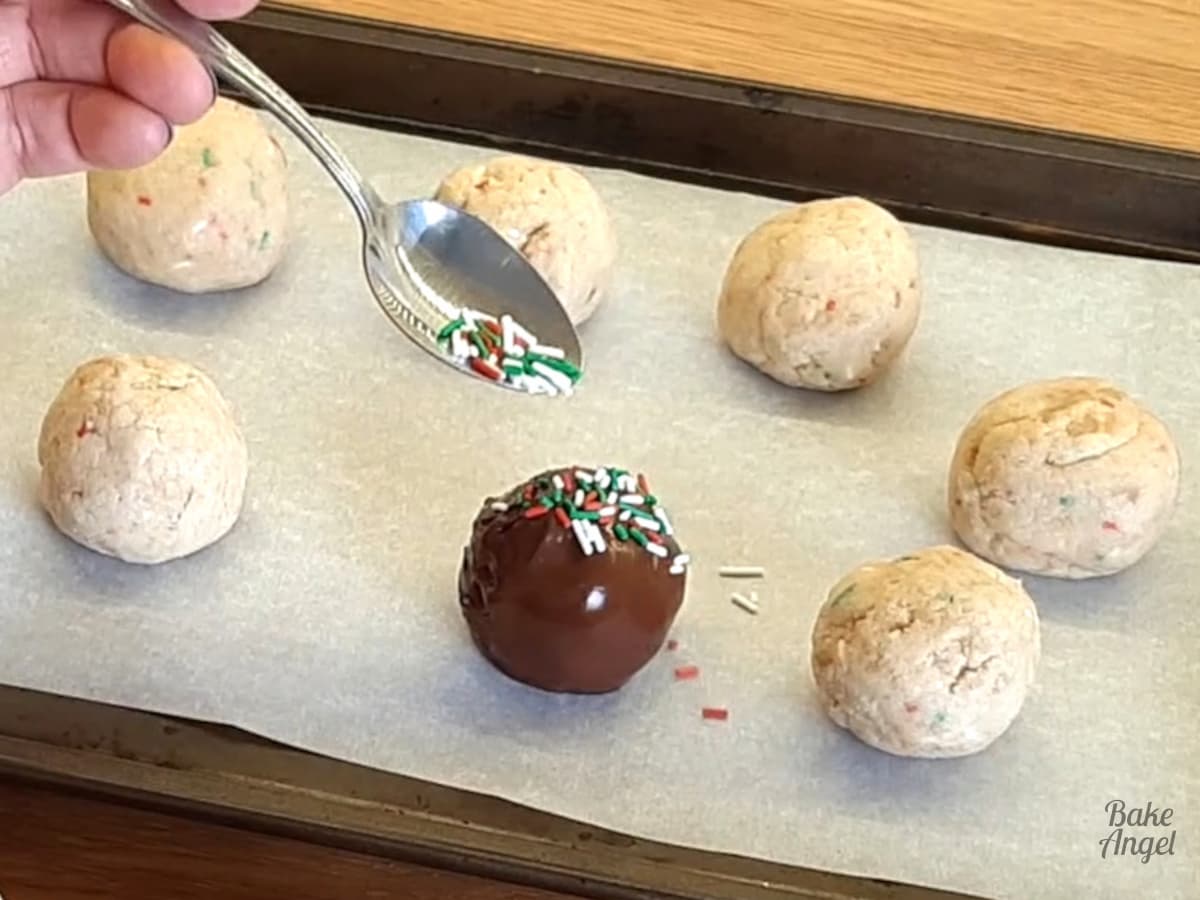 Cake balls on a lined baking tray. Sprinkles are being added to a freshly chocolate coated cake ball. 