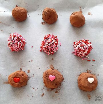9 oat cocoat truffles on parchment paper in rows of 3. The top row is coated in cocoa powder, the middle row is coated in pink, red and white sprinkles and the botton row is coated in cocoa with a candy heart in the middle.