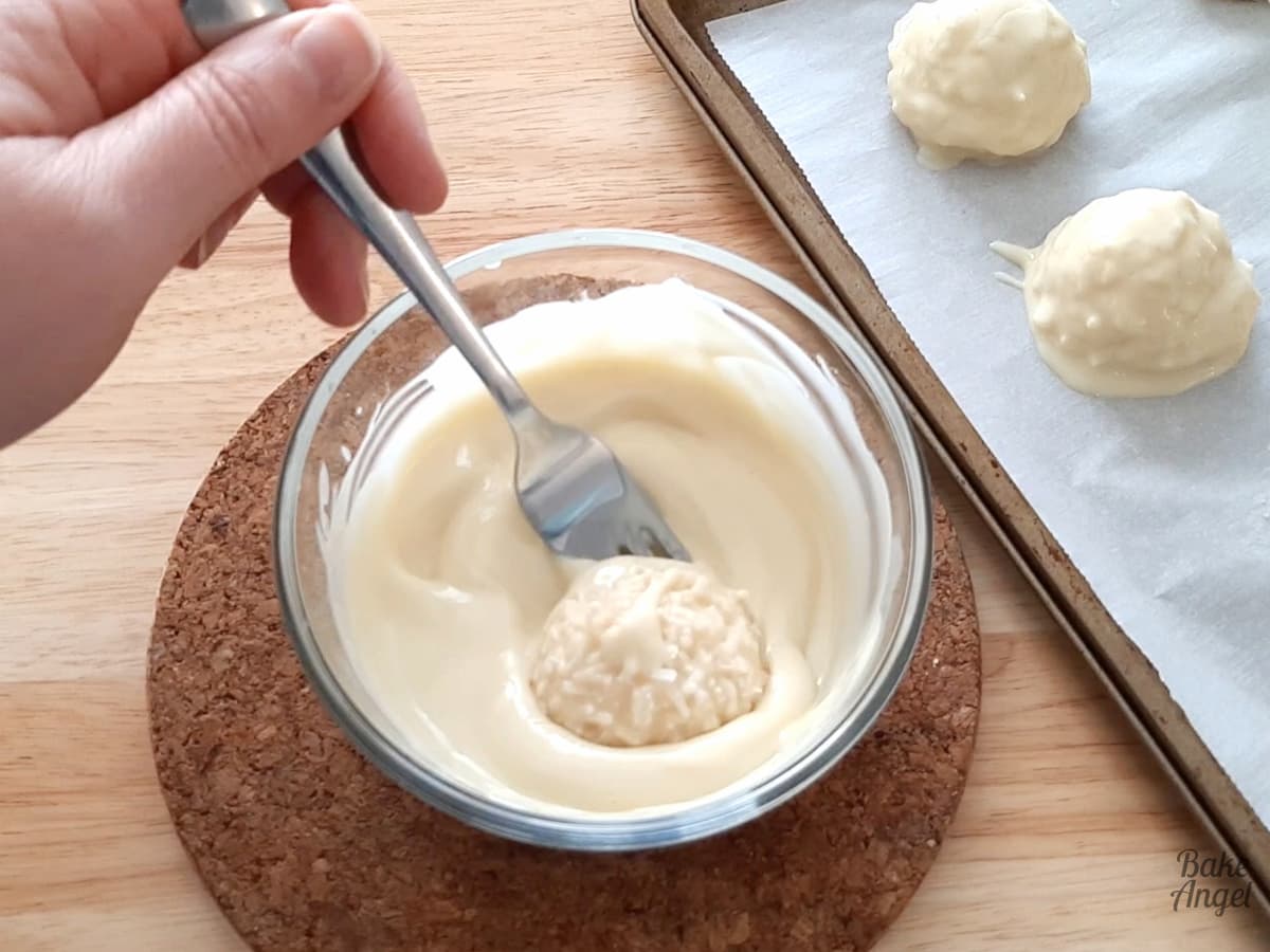 A coconut bliss ball being dipped in melted white chocolate and using a fork to turn it over to coat it.. 