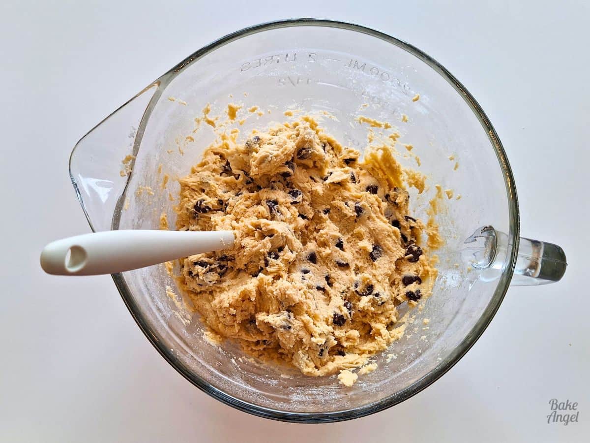 A glass mixing bowl containing chocolate chip cookie dough and a cream coloured silicone spatula.