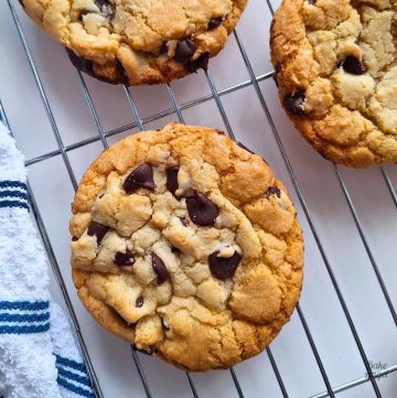 Close up of three copycat Disney chocolate chip cookies on a cooling rack. There is part of a white and blue tea towel on the left of the rack.