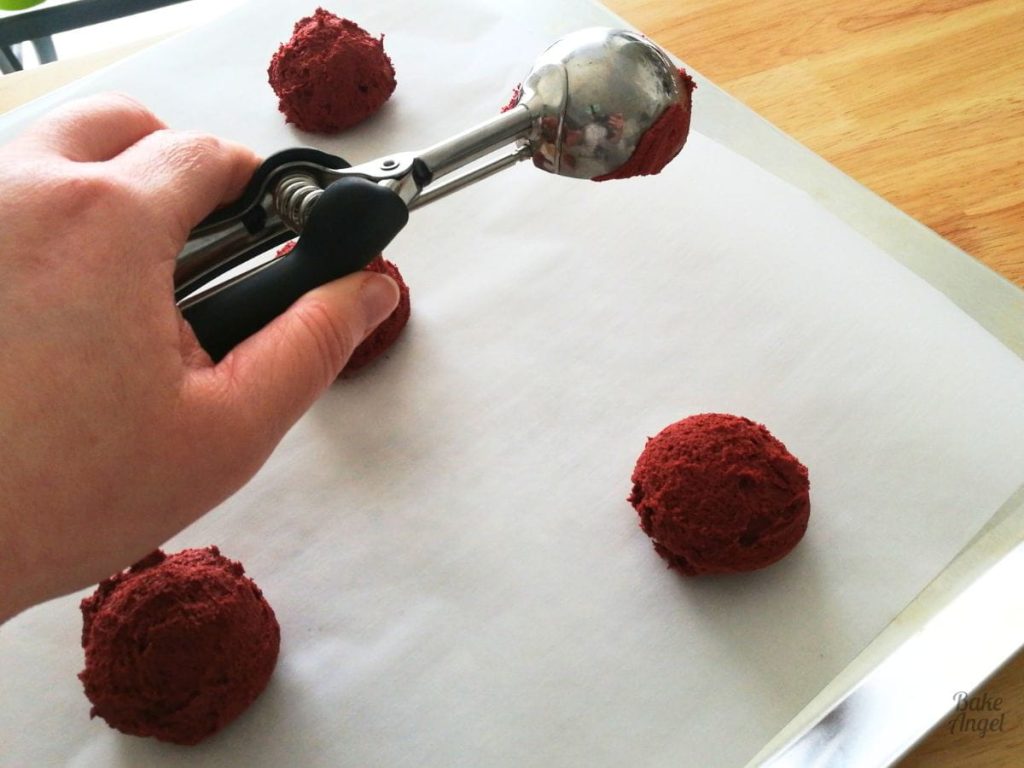 Balls of red velvet cookie dough being scooped on to parchment paper using a cookie scoop.