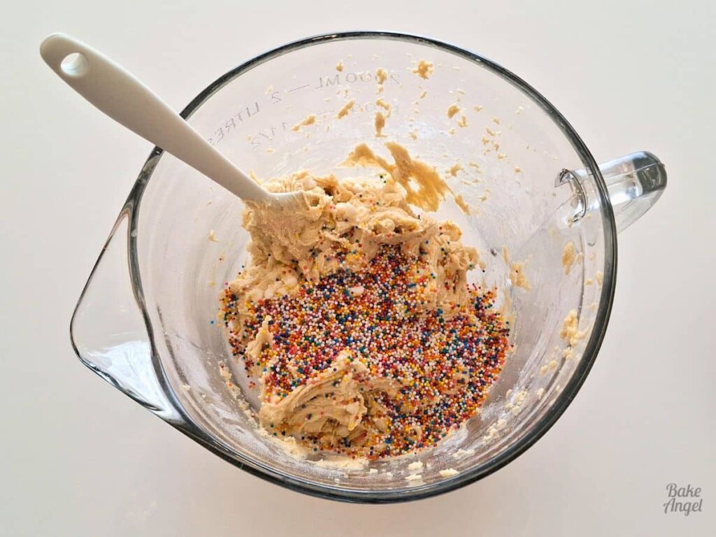 Rainbow non-pareils on top of cookie dough in a mixing bowl.