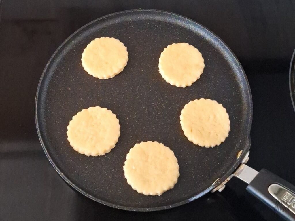 5 welsh cakes baking on a round griddle pan on a stovetop.