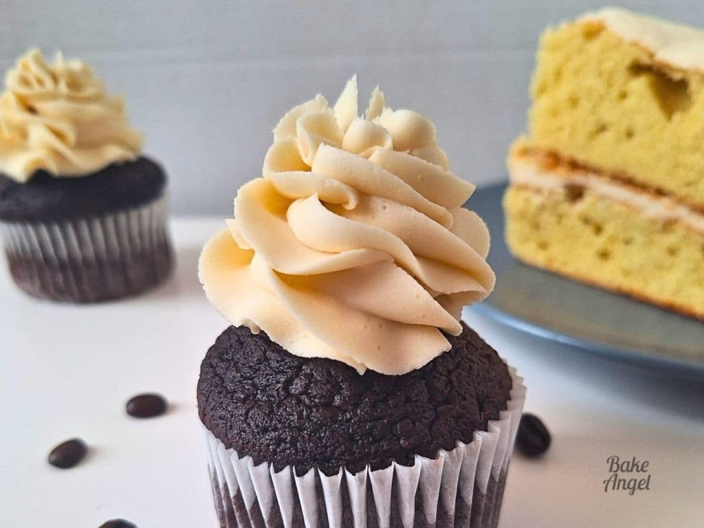 A wide shot of Vegan Coffee Buttercream Frosting swirled on a chocolate cupcake. There is a slice of coffee layer cake and another cupcake in the background.