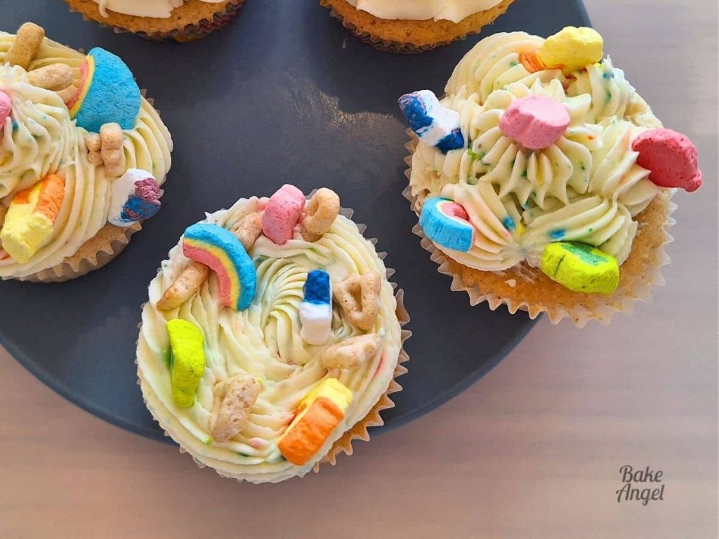 Cupcakes on a plate decorated with Lucky Charms Buttercream swirls, marshmallows on cereal pieces.