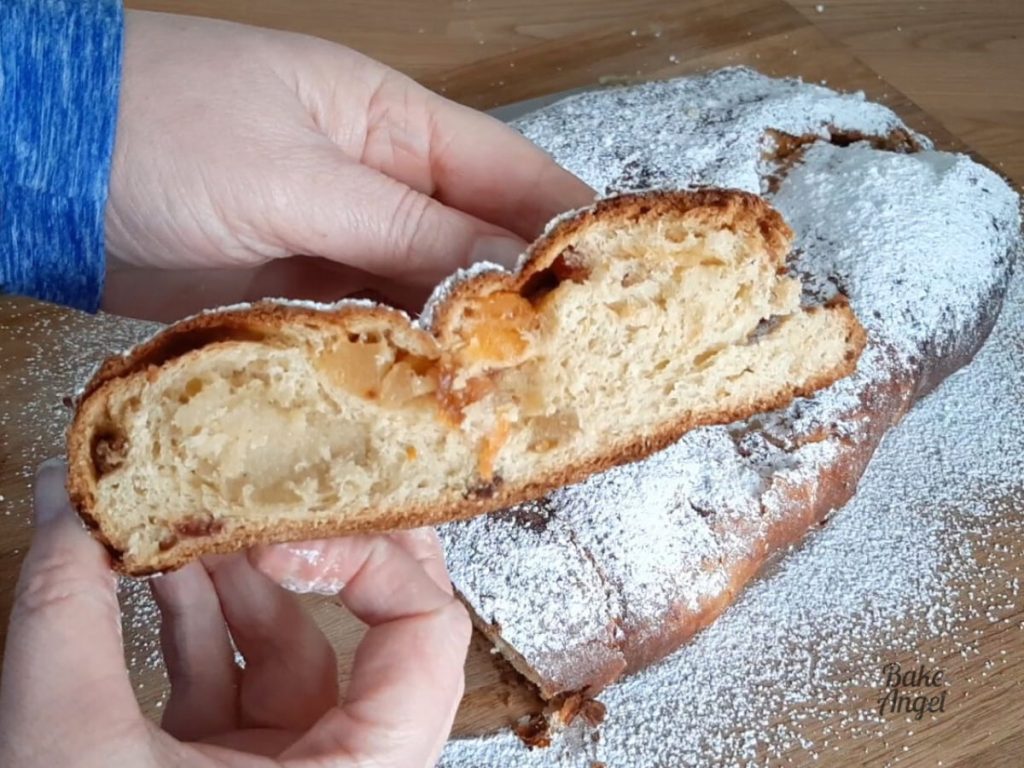 Close up of a slice of stollen being held over a wooden board. The slice shows a ribbon of marzipan and dried fruit throughout.