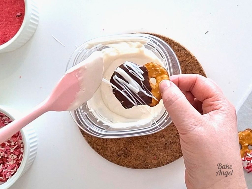 Drizzling white chocolate from a pink spatula on to a chocolate dipped pretzel.