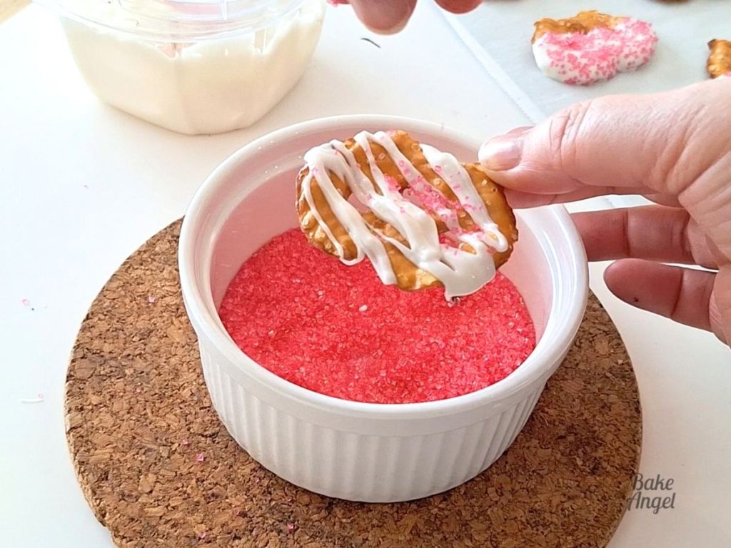 Adding pink sanding sugar to a white chocolate drizzle pretzel.