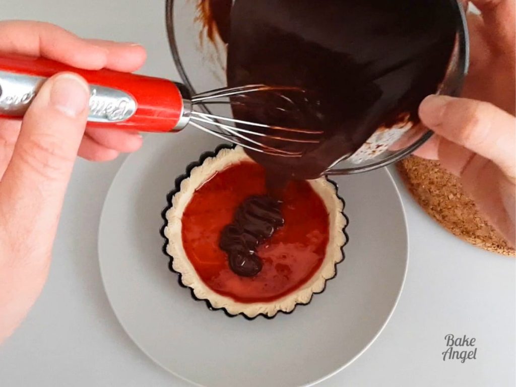 Chocolate Ganache being poured onto a mini tart filled with strawberry jam.
