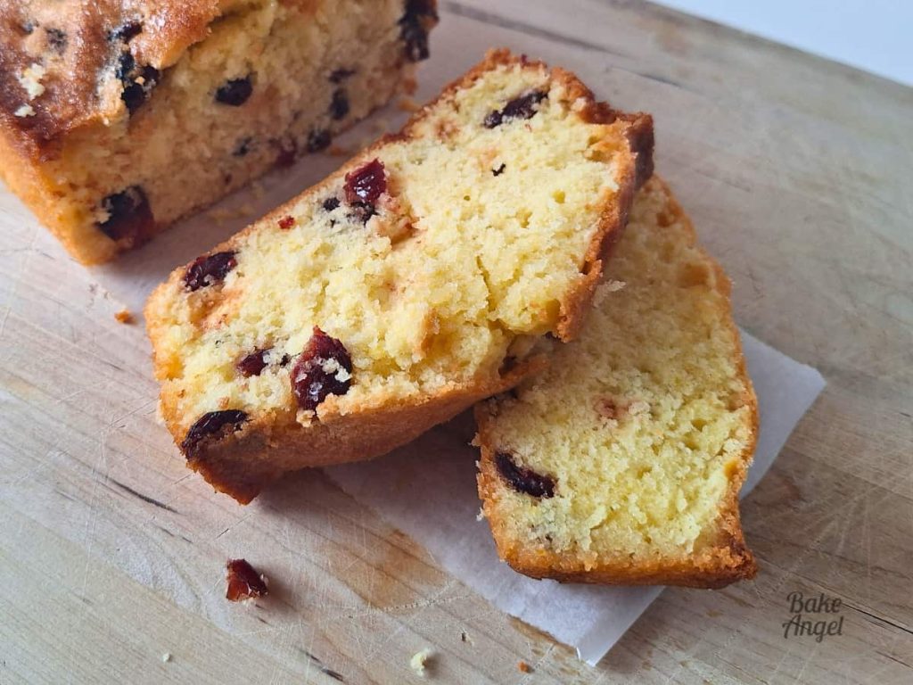 Two slices of dairy free lemon cranberry loaf cake on a wooden board. The slices have pops of cranberries in them.