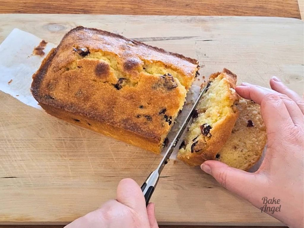 A hand holding a slice of lemon cranberry loaf cake while another slice is being cut.