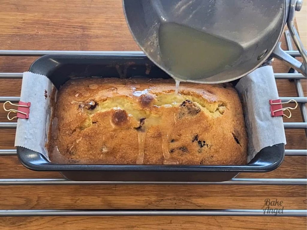 Lemon glaze being poured over a warm lemon cranberry loaf cake in a pan.