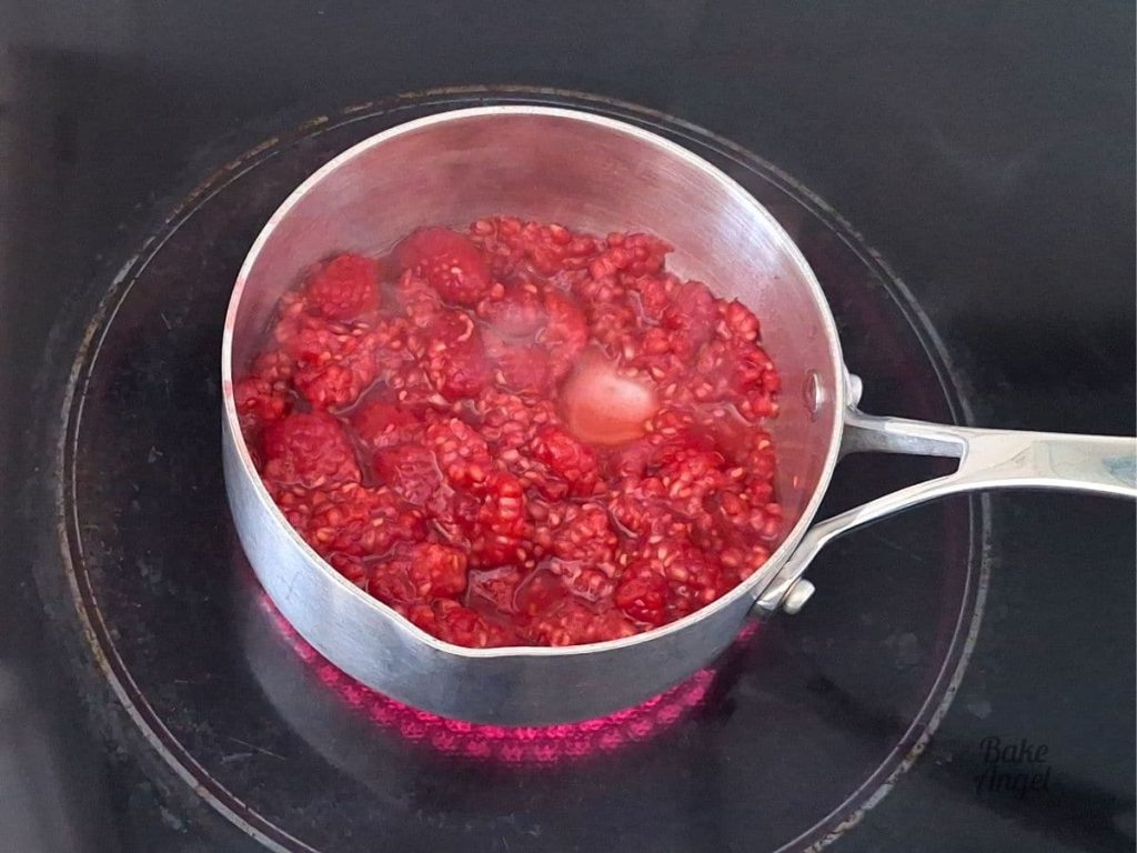 Close up of raspberries, water and sugar being cooked in small saucepan on a stovetop.