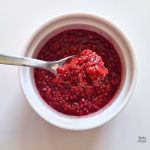 Close up of a small batch of 3 ingredient raspberry compote in a white ramekin, with a small amount on a spoon being held over the ramekin.
