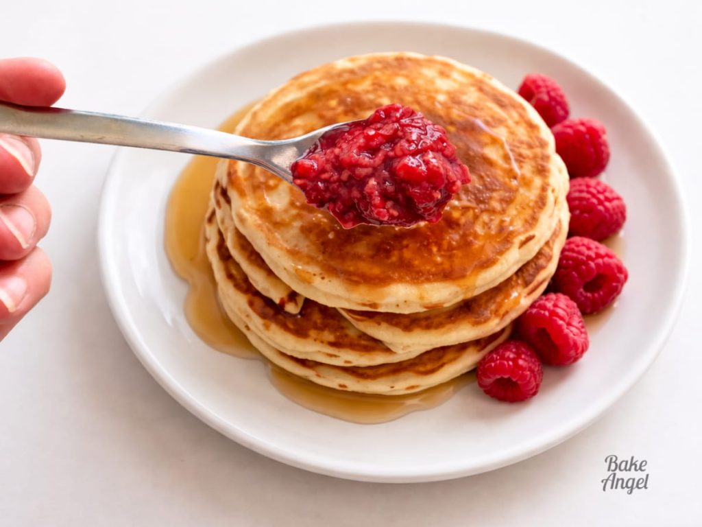Close up of a spoonful of 3 ingredient raspberry compote held over a plate of stacked pancakes, maple syrup and fresh raspberries.