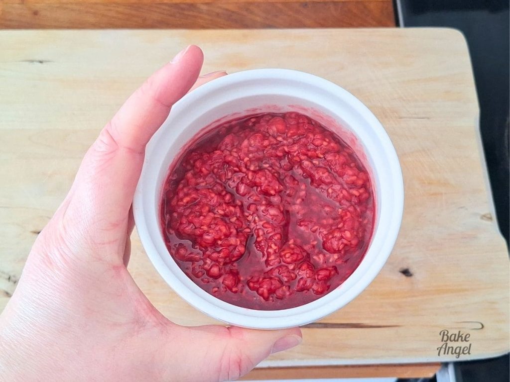 A small white ramekin filled with raspberry compote, being held over a wooden board.