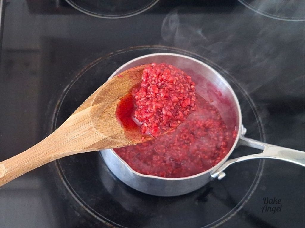 Raspberry compote on a wooden spoon, being held over a saucepan on the stovetop.