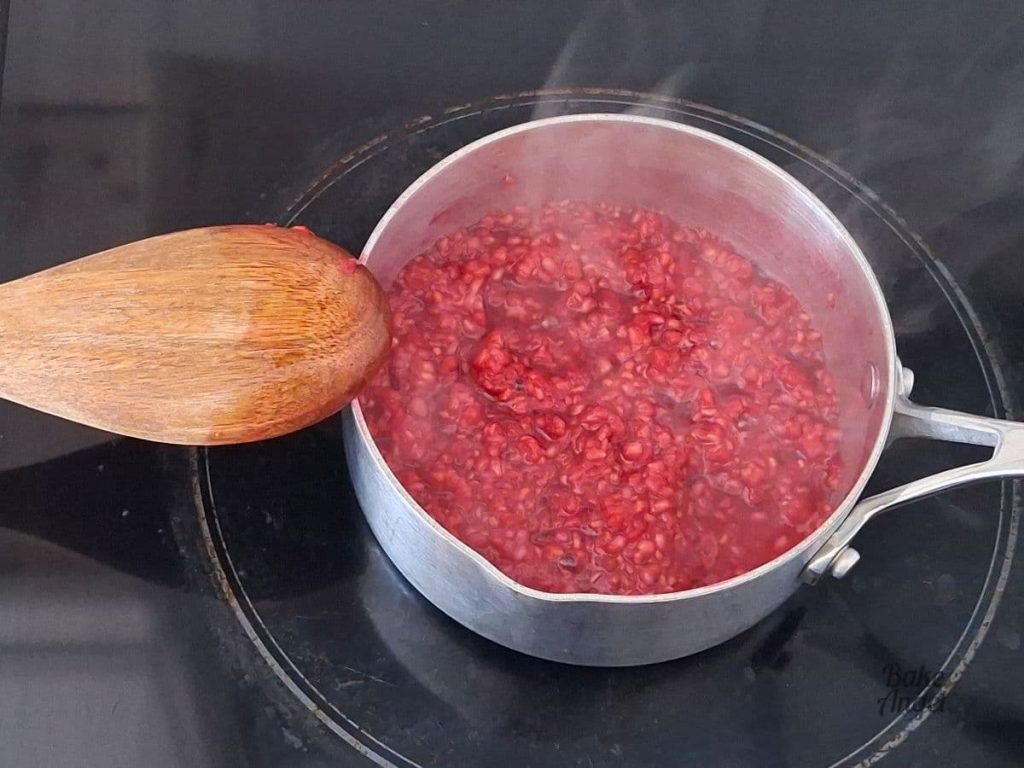Raspberry compote cooking in a small saucepan on a stovetop -there is visible steam coming from the saucepan.