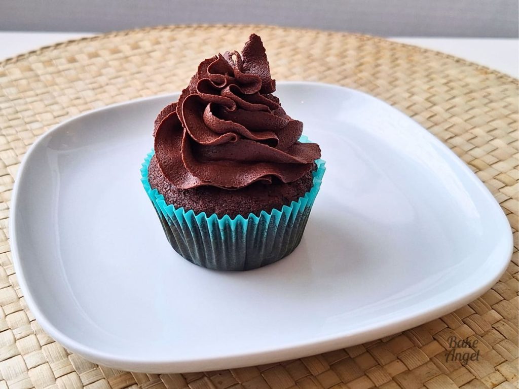 A dairy free cupcake decorated with chocolate buttercream frosting on a white plate. The cupcake is in a teal coloured cupcake liner.