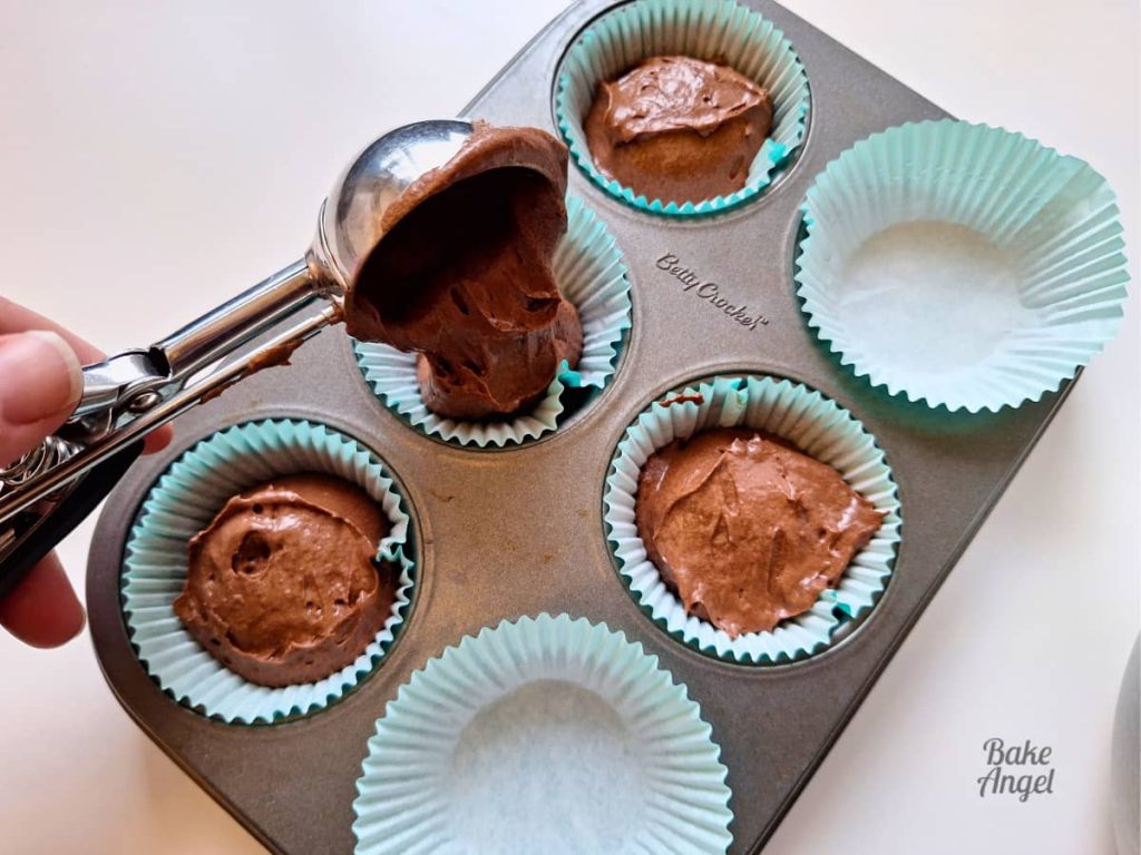 An ice cream scoop being used to add a scoop of batter to a teal cupcake liner. The liners are in a 6 cavity cupcake baking pan.