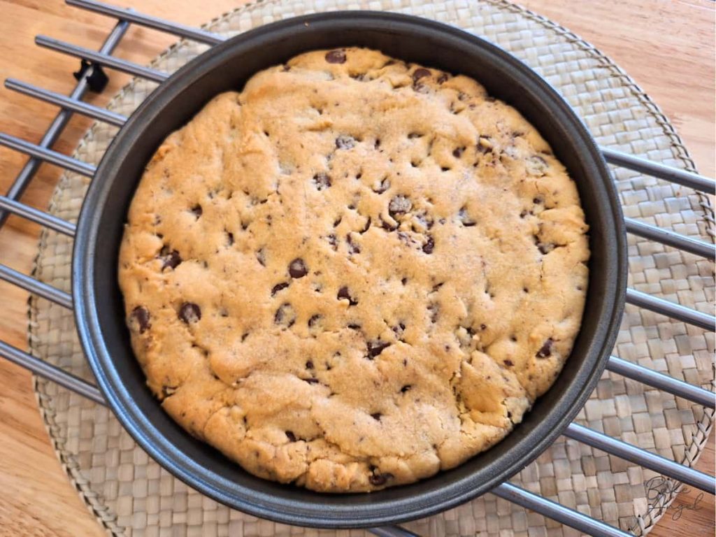 A round cake pan filled with a chocolate chip cookie cake, on a cooling rack.