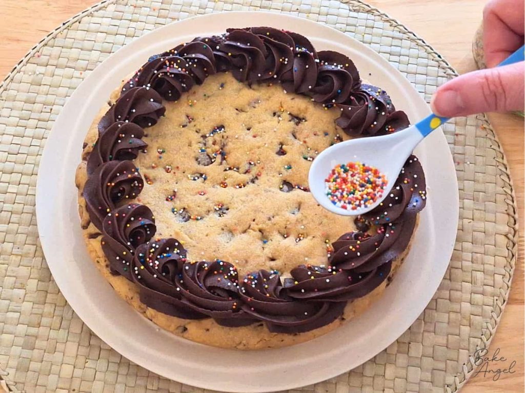 Sprinkles being added to a dairy free chocolate chip cookie cake, decorated with chocolate buttercream.