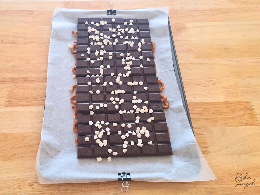 White chocolate chips on top of 4 bars of dark chocolate and pretzels for making vegan pomegranate pretzel bark.