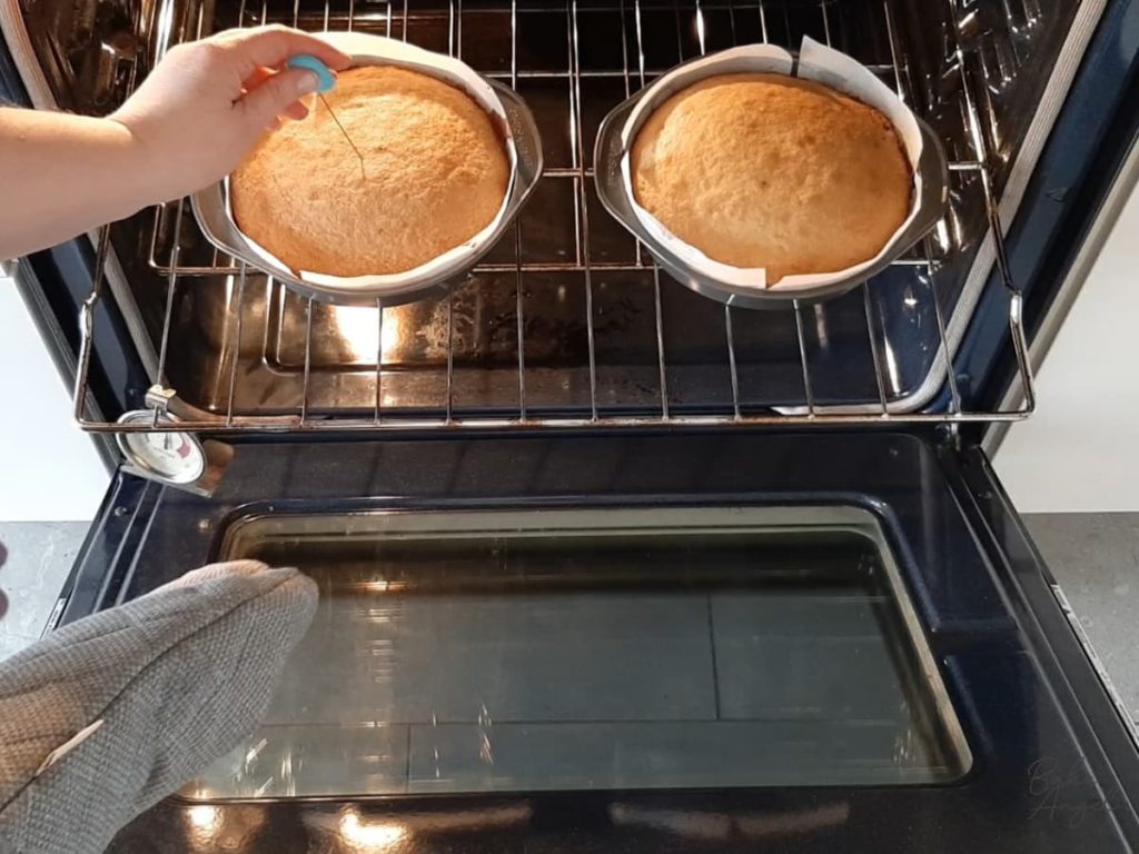 Two baked cakes on an oven shelf, with one being tested for doneness with a cake tested.