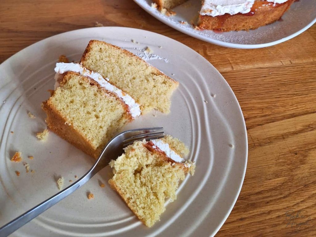 A slice of Victoria sponge cake on a plate with a fork cutting it.