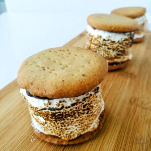 Three S'mores on a wooden board - each s'more has two digestive biscuits, chocolate ganache and a toasted marshmallow in the middle.