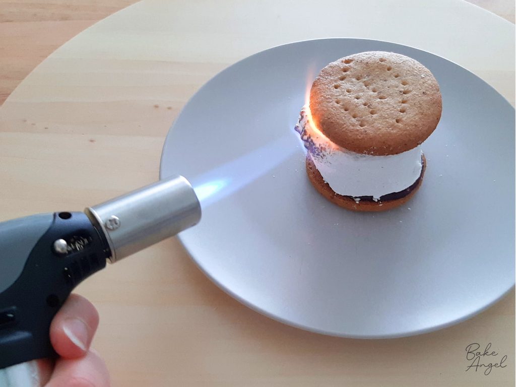 A mini kitchen blowtorch being used to torch a marshmallow in a s'more. The flame is visible on the s'more.
